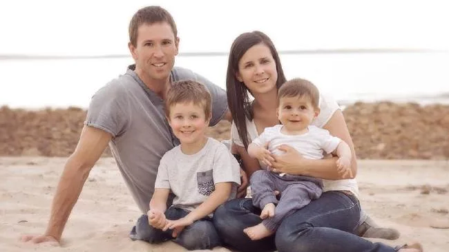 A family of four sitting on the beach with a serene ocean backdrop, parents holding two young children, all smiling.
