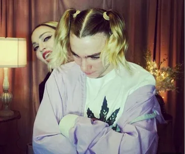 Two individuals with playful expressions, one facing the camera and one looking down, in a warmly lit room.