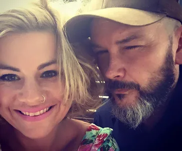 Two people close together; one smiling with a nose ring, the other with a beard and cap, both in casual attire.