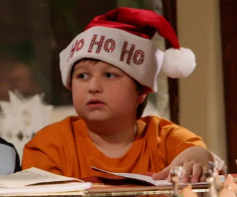 Boy in an orange shirt wearing a Santa hat with "Ho Ho Ho," sitting at a table with papers.
