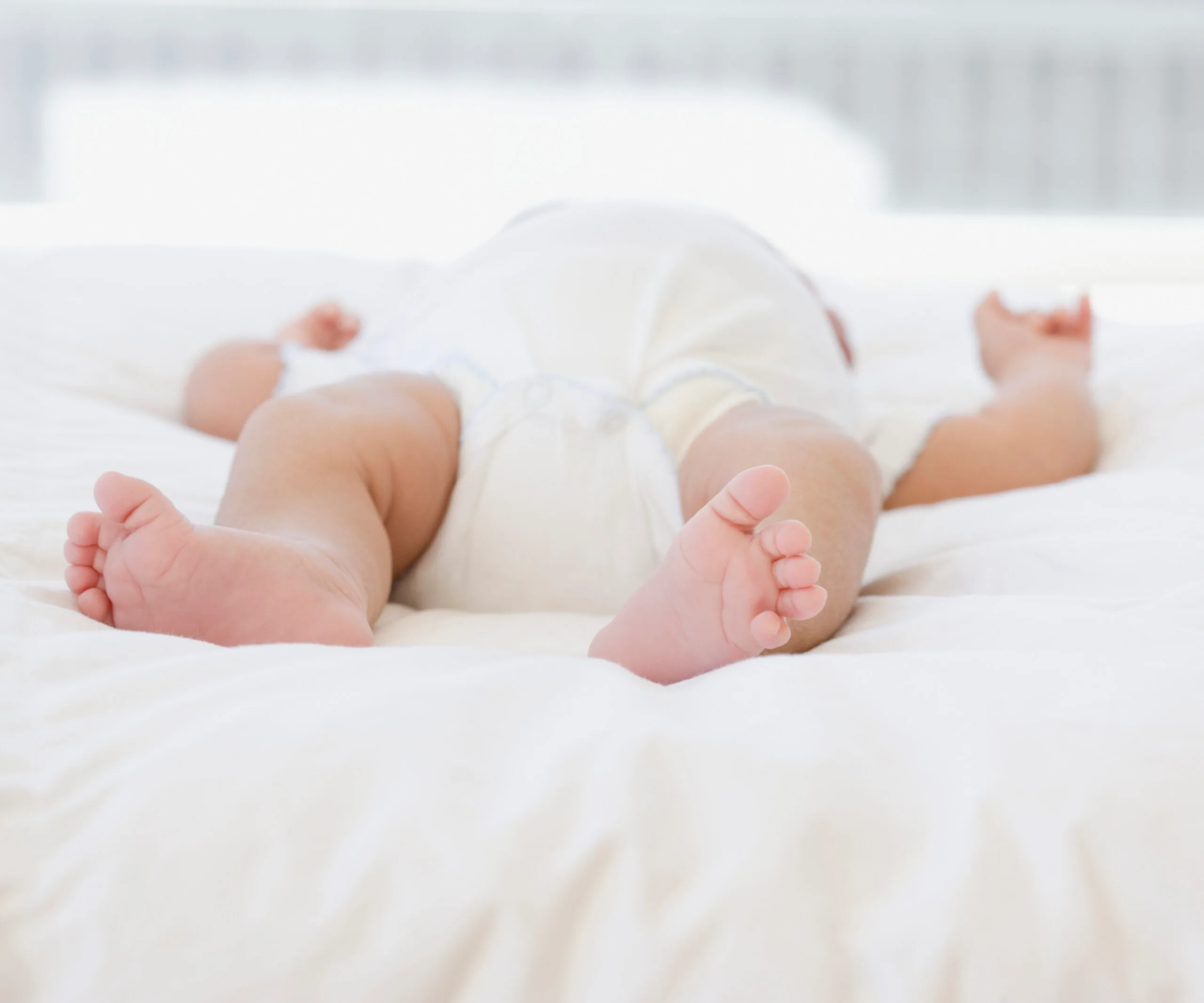 A baby lying on a soft, white bed, wearing a diaper, viewed from the feet upward.