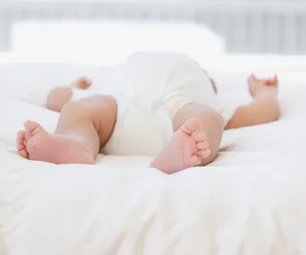 A baby lying on a soft, white bed, wearing a diaper, viewed from the feet upward.
