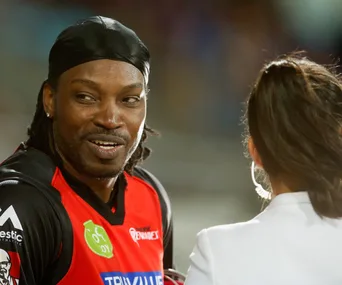 A cricketer in a red jersey smiles while talking to a woman in a stadium setting.