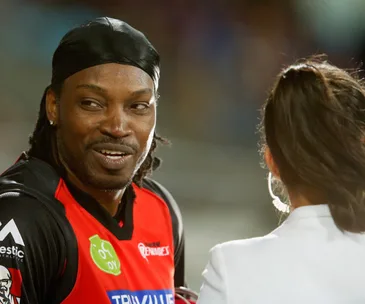 A cricketer in a red jersey smiles while talking to a woman in a stadium setting.