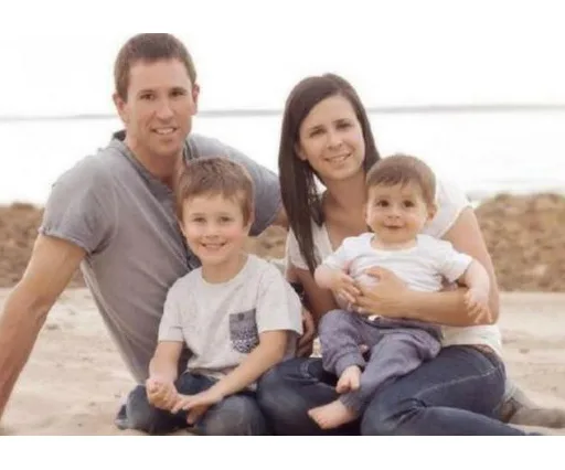 Family sitting on a beach, smiling; two adults and two young children, one sitting on the mother's lap.