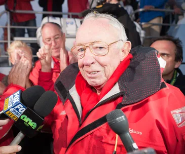Man in red jacket interviewed by media, surrounded by applauding crowd.