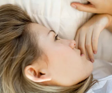A woman lying down on a bed, looking contemplative, resting her head on a pillow with a neutral expression.