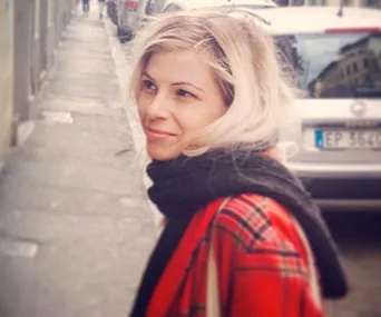 A woman with blonde hair wearing a red plaid coat and black scarf stands on a city sidewalk, smiling slightly.