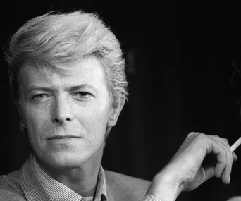 A person with styled hair, holding a cigarette, gazing thoughtfully, black and white image.