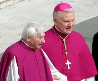 Two clergymen in ceremonial attire, one with a pink zucchetto and cross necklace, walking together.
