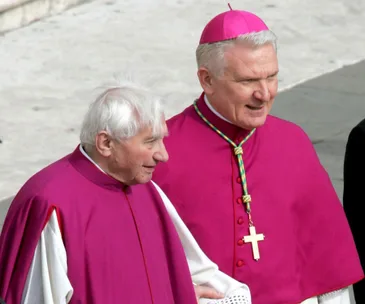 Two clergymen in ceremonial attire, one with a pink zucchetto and cross necklace, walking together.