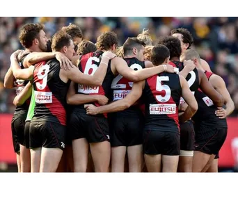 Essendon Bombers players huddle in black and red uniforms during a match, displaying team unity.