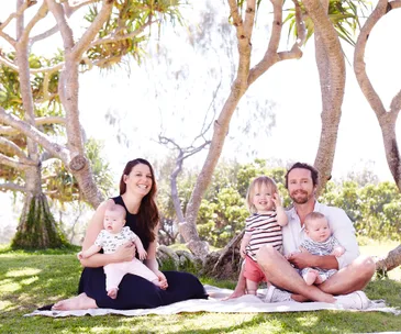 A happy family with two babies and a toddler sitting on a picnic blanket under trees in a sunny park.