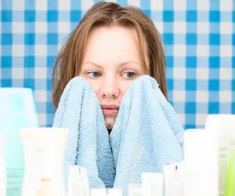 "Person holding a blue towel to face, surrounded by toiletries, with a blue checkered background."
