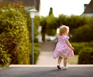 Toddler in a floral dress walks alone on a path surrounded by greenery.