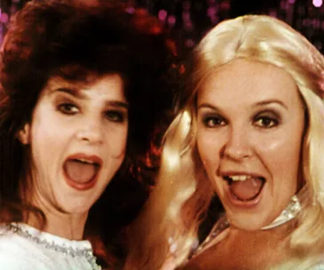 Two women with open mouths smiling, dressed colorfully, from a scene reminiscent of the film "Muriel's Wedding."