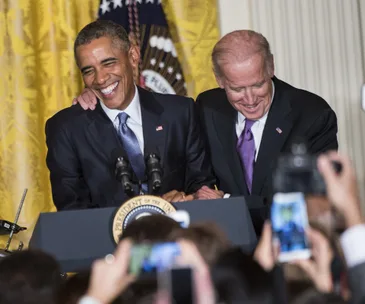 Two men laughing and embracing at a podium with microphones, taken in a formal setting with audience capturing the moment.