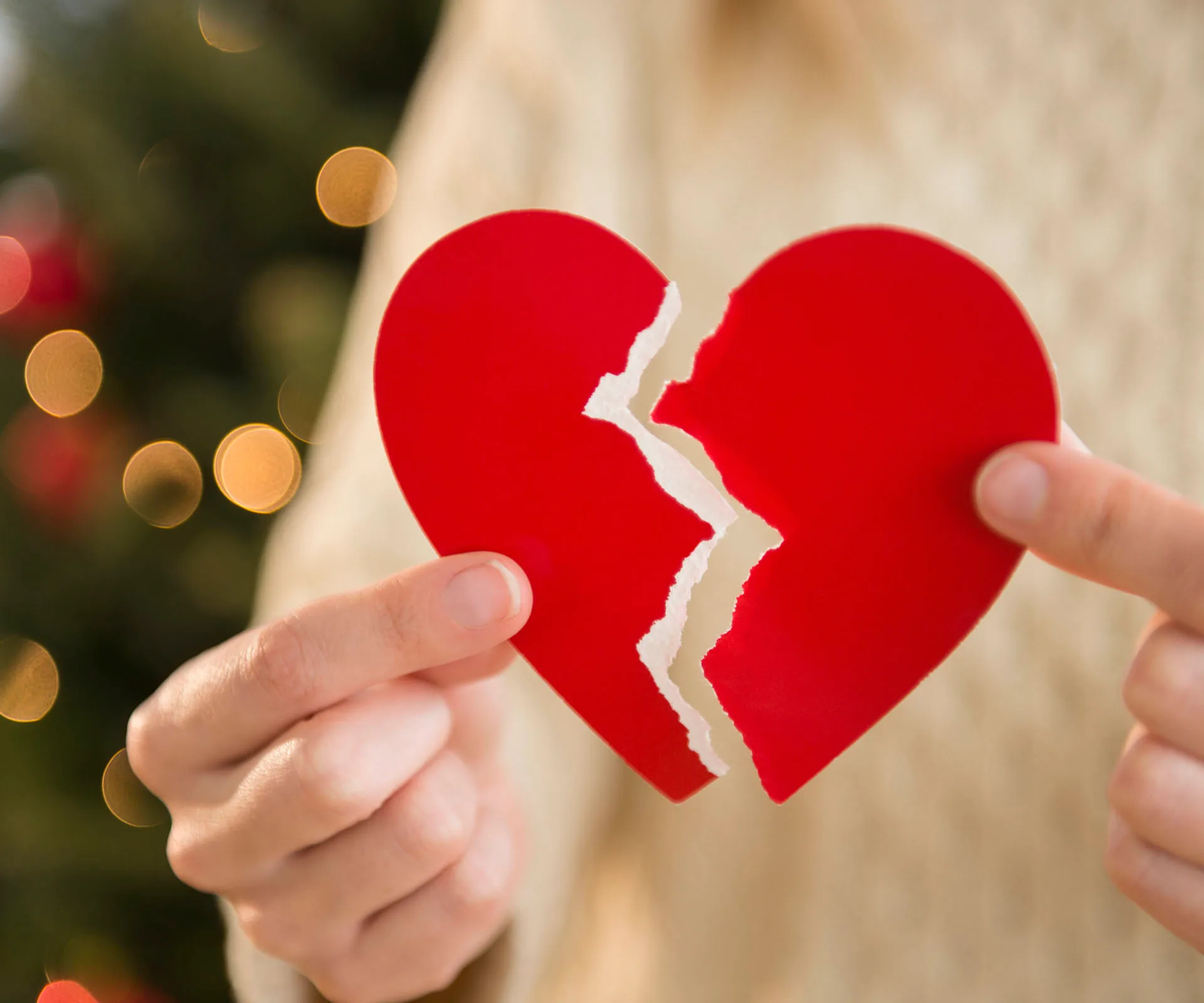 A person holding two halves of a torn red paper heart against a blurred background.