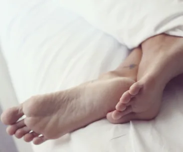 Bare feet with a small tattoo relax under a white sheet, highlighting a serene bedroom scene.