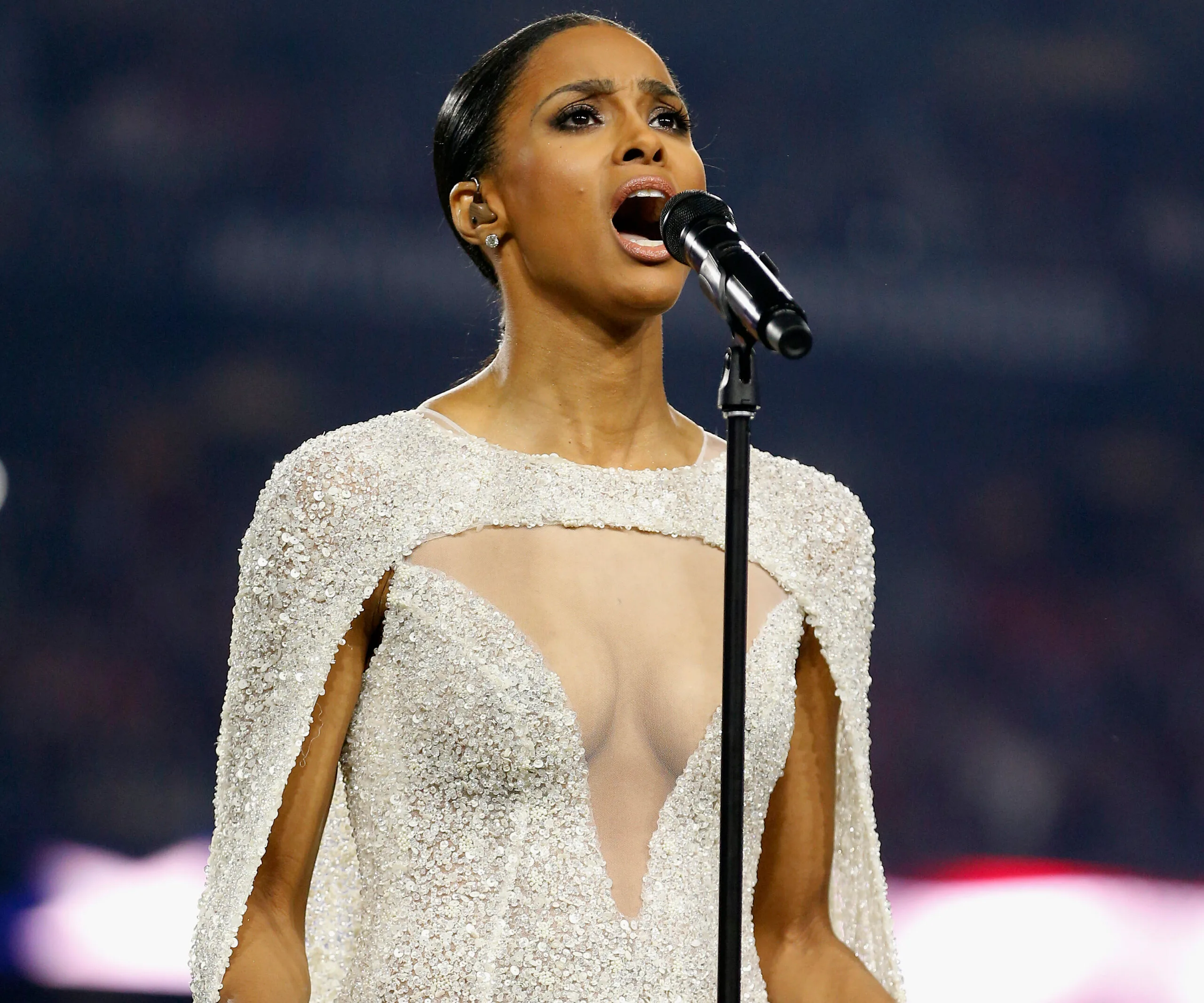Singer performing the national anthem in a sparkling, low-cut white dress on stage.