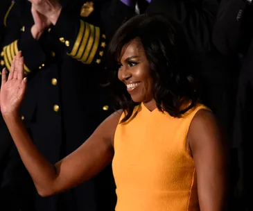 Michelle Obama waves, wearing a sleeveless yellow Narciso Rodriguez dress at an event.