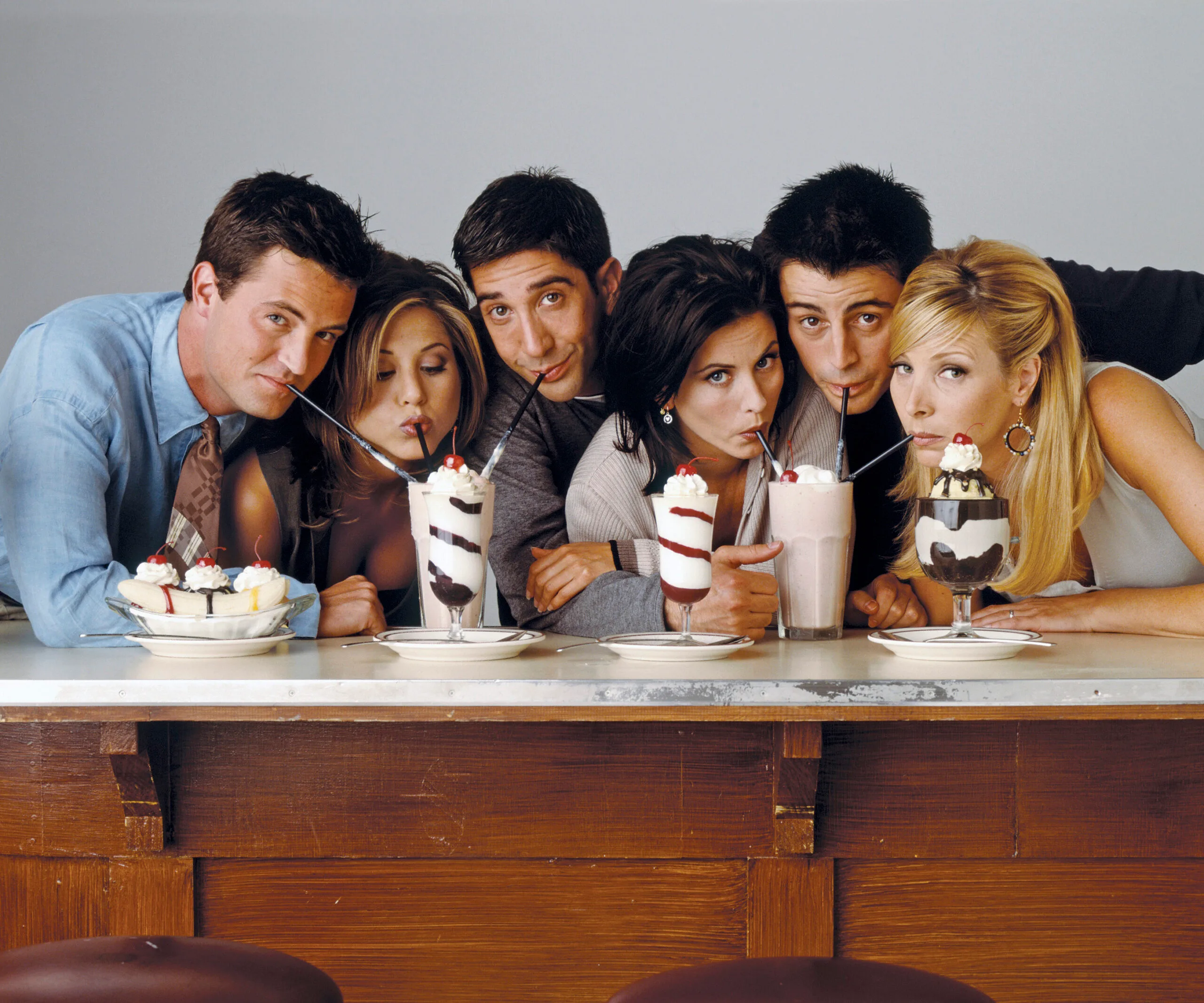 Cast of "Friends" sipping milkshakes at a diner counter, all leaning close together.
