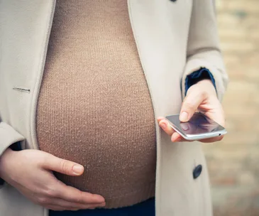 Pregnant woman in a coat holding her belly with one hand and a smartphone in the other.