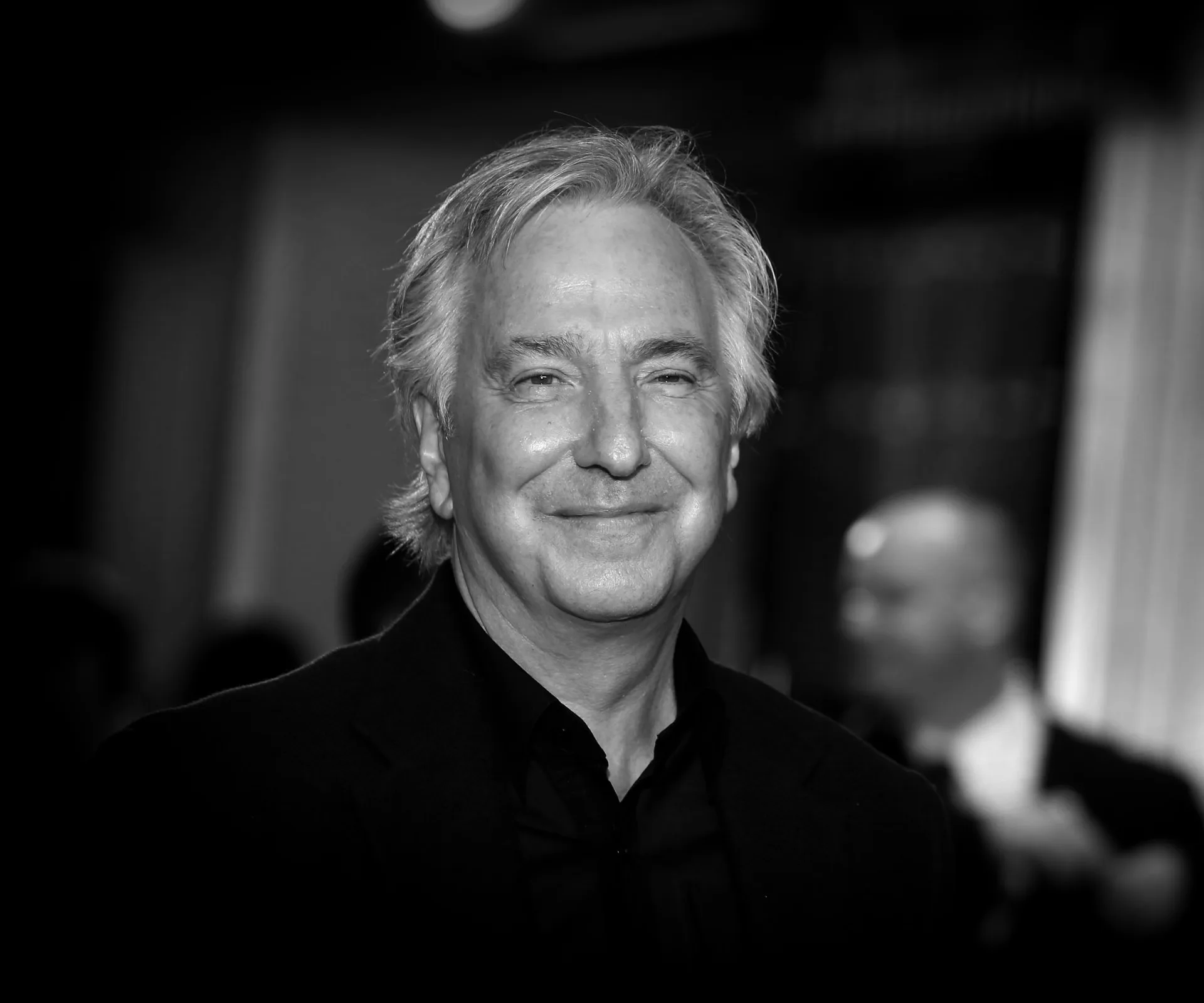 A black and white portrait of Alan Rickman, smiling in a suit.