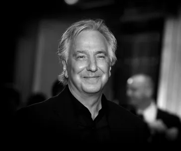 A black and white portrait of Alan Rickman, smiling in a suit.