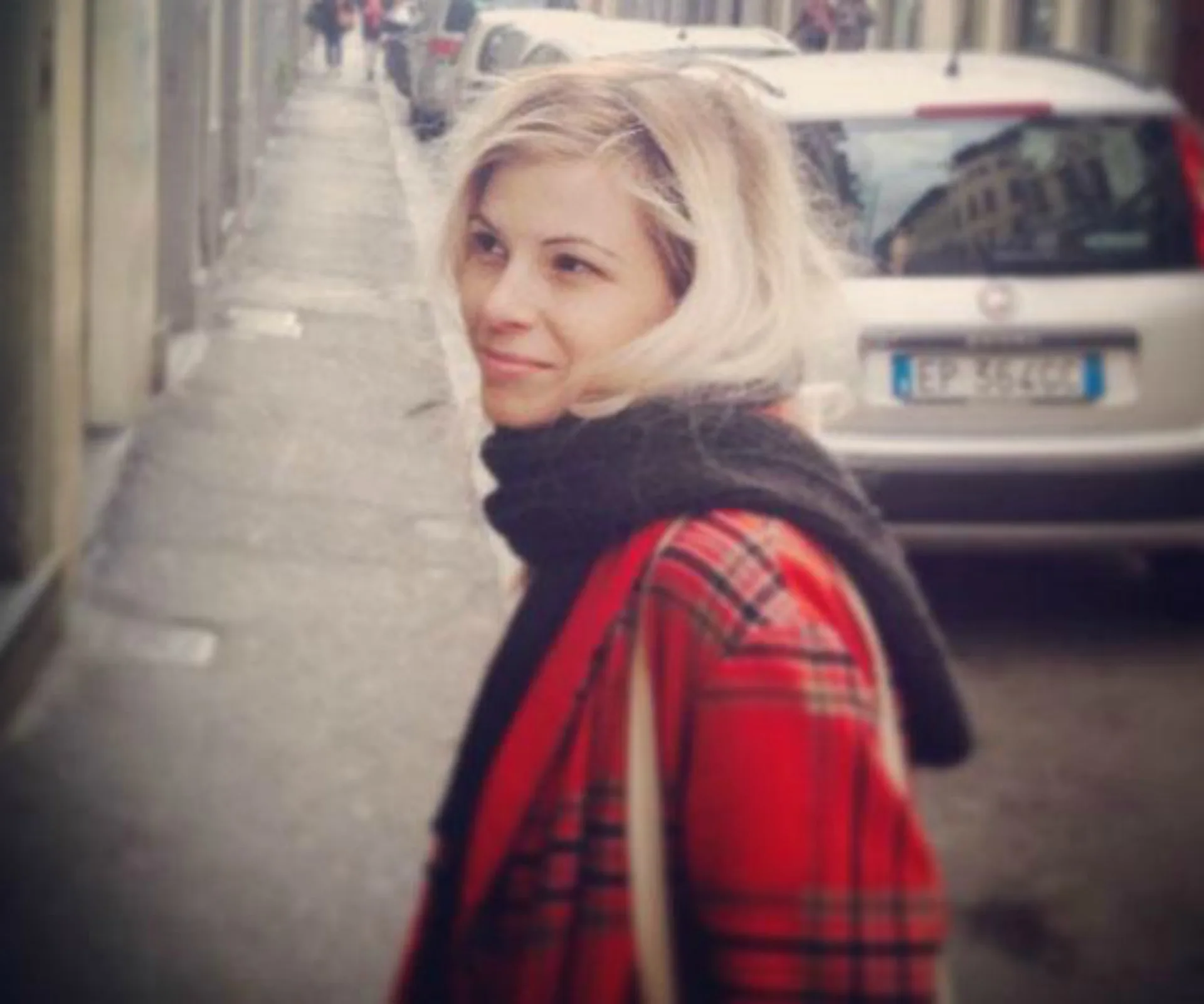 A woman with blonde hair and a red plaid coat smiles while standing on a street with parked cars.