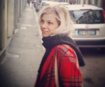 A woman with blonde hair and a red plaid coat smiles while standing on a street with parked cars.