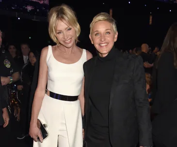 Two smiling women, one in a white dress and the other in a black outfit, at an event with a crowd in the background.