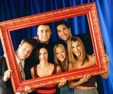 A group of six friends smiling and holding a large red frame, set against a blue background.