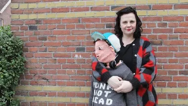 Woman holding a knitted replica of her son against a brick wall, both dressed in casual attire.
