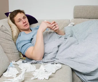 A man lying on a couch, appearing sick, covered with a blanket, surrounded by tissues.
