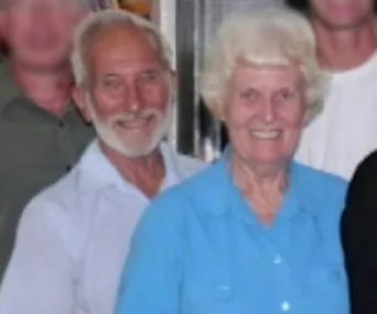Elderly couple smiling together indoors; man with white beard and woman in blue shirt.