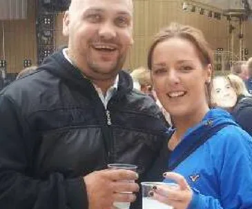 A smiling man and woman holding drinks, standing closely in an outdoor setting.