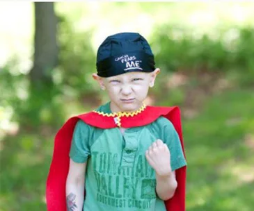 A young child wearing a red cape and black hat stands outdoors, with a determined expression, t-shirt reading "Cancer Fears Me".