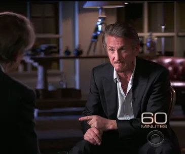 Actor in suit during "60 Minutes" interview, gesturing with one hand inside a dimly lit room.