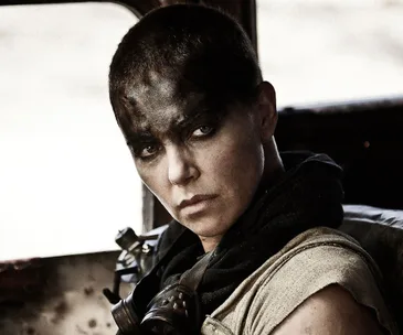 A woman with short hair and black face paint looks intently from a vehicle in a post-apocalyptic setting.