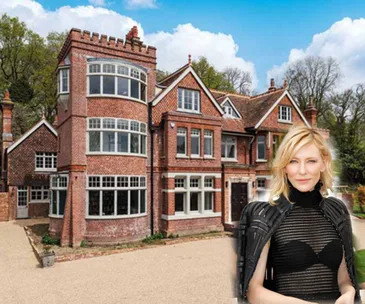 Cate Blanchett in front of a large, brick English manor with gabled roof and tower under a blue sky.