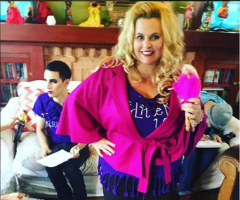 A woman in a pink outfit holds a Barbie doll, standing in a living room with others seated in the background.