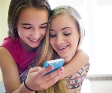 Two girls smiling and looking at a smartphone together. One has braces and both are enjoying the moment.