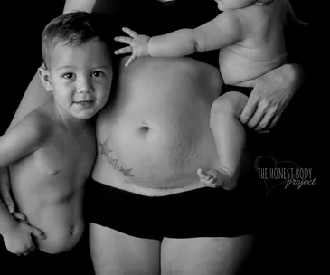 Mother holding baby with toddler hugging her bare postpartum belly, showing stretch marks and star tattoo.
