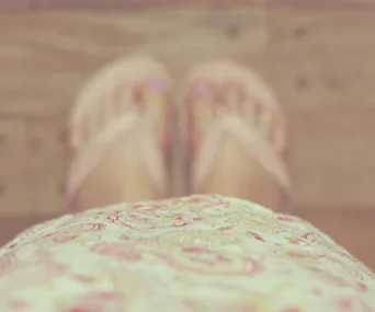 Blurry view looking down at feet in flip-flops and floral dress on wooden floor.