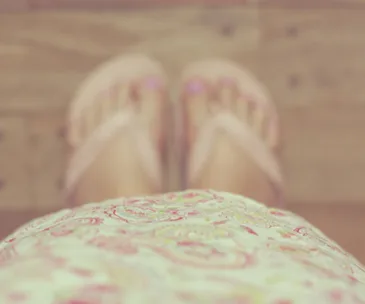 Blurry view looking down at feet in flip-flops and floral dress on wooden floor.