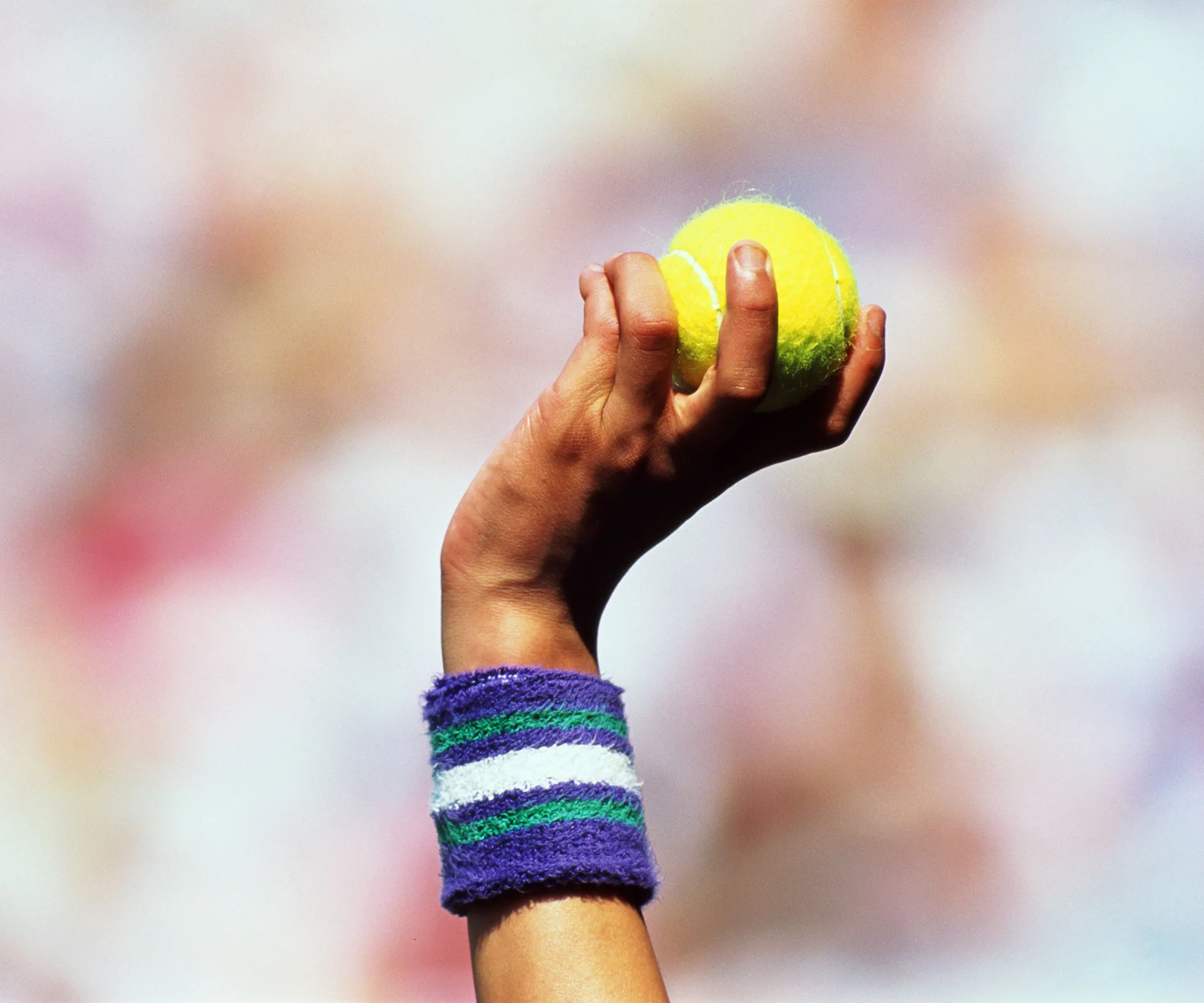 A hand with a striped wristband holding a yellow tennis ball against a blurred background.