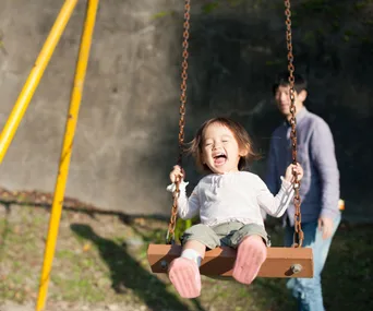 Child joyfully swinging, pushed by an adult in a park; sunny day with a blurred background.