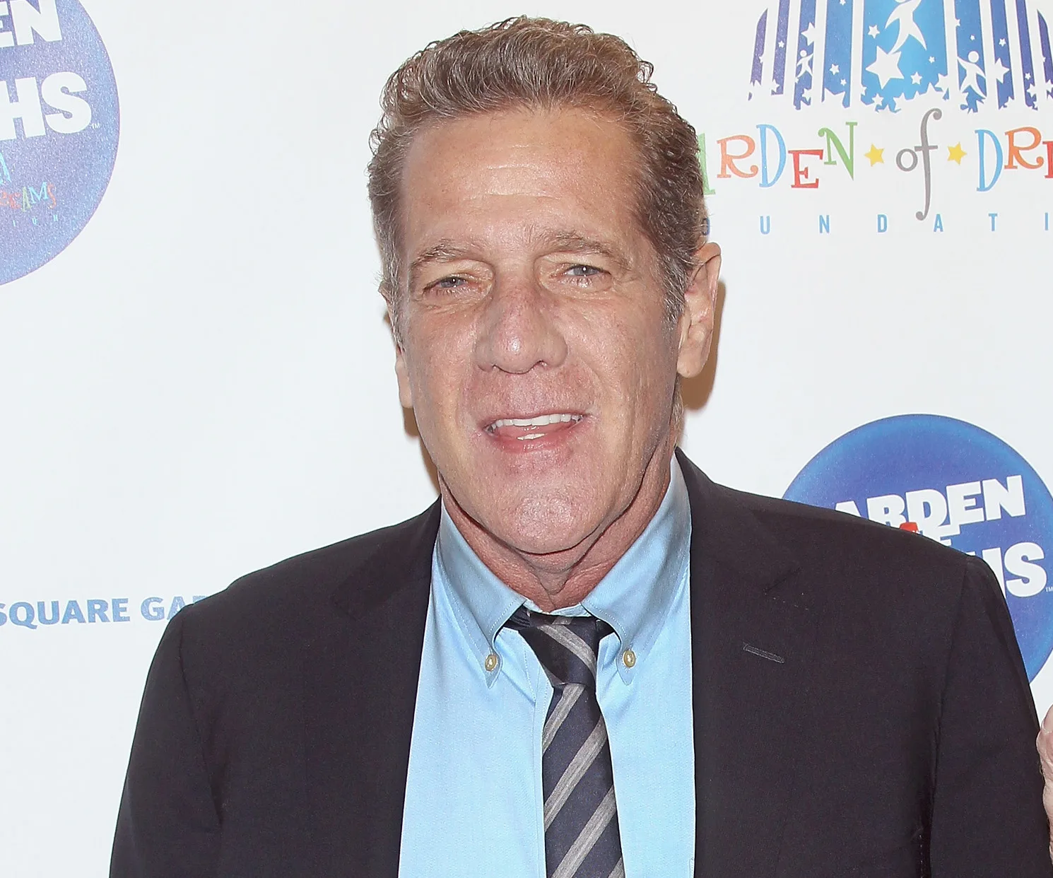 A man in a suit and tie smiles at an event with a "Garden of Dreams" backdrop.