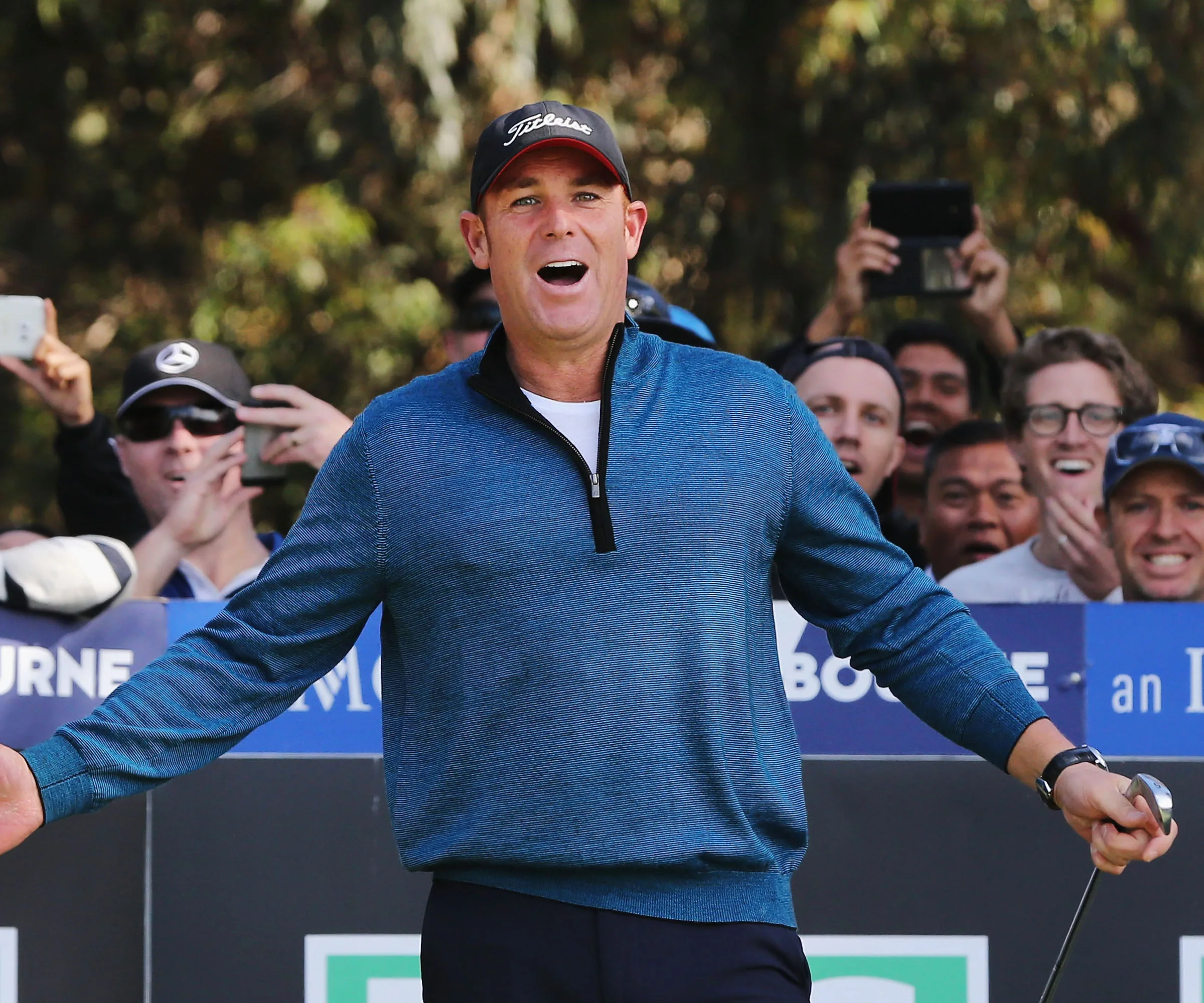 Shane Warne at a golf event, excitedly reacting with a club in hand, surrounded by a cheering crowd.